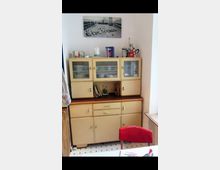 Ein Retro-Küchenschrank aus Holz mit cremefarbenen Türen und Glaseinsätzen steht an einer weißen Wand. Darüber hängt ein schwarz-weißes Foto, auf dem eine städtische Szene abgebildet ist, und es befinden sich diverse Küchengeräte und Dekorationsgegenstände auf der Schrankoberfläche.