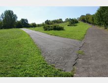 Ein weitläufiger Park mit zwei gepflasterten Wegen, die sich gabeln und von grüner Wiese umgeben sind. Im Hintergrund sind Bäume und Büsche sowie sanfte Hügel sichtbar.