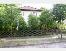 Zweistöckiges Wohnhaus mit weißer Fassade und rotem Ziegeldach, umgeben von Bäumen und Sträuchern. Vor dem Grundstück verläuft ein Gehweg, der durch einen Holzzaun vom Garten getrennt ist, und daneben liegt eine gepflasterte Straße.