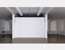 Leerer, heller Raum mit glatten Böden, einer weißen Trennwand in der Mitte und sichtbaren Rohrleitungen an der Decke. An den Seiten befinden sich große, helle Fenster mit blickdichten Rollläden sowie zwei Feuerlöscher in einer Ecke.