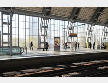 Bahnhof Alexanderplatz in Berlin mit modernem Bahnsteig und großer Fensterfront, die viel Tageslicht hereinlässt. Mehrere Personen stehen auf dem Bahnsteig, einige nahe einer Rolltreppe und einem Werbeplakat.