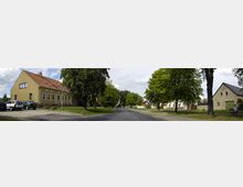 Straßenszene in einem Dorf mit einer asphaltierten Straße, gesäumt von grünen Bäumen. Links befindet sich ein gelbes Gebäude mit roten Dachziegeln und parkenden Autos, rechts ein älteres Gebäude mit einem Giebeldach.