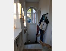 Heller Eingangsbereich mit einer weißen, halbhohen Tür und darüber einem großen, bogenförmigen Fenster, das Tageslicht hereinlässt. Links befinden sich Regale mit Ablageflächen, rechts hängen Taschen und Kleidungsstücke an Haken; ein kleiner Hund steht auf einem Läufer und blickt zur Tür.