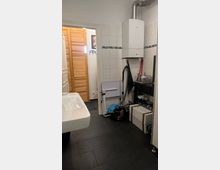 Badezimmer mit weißen Fliesen und dunklem Boden, links ein Waschbecken, rechts ein weißer Wandboiler, ein Regal mit Gegenständen und ein zusammenklappbarer Duschsitz; im Hintergrund eine Tür mit Holzlamellen.