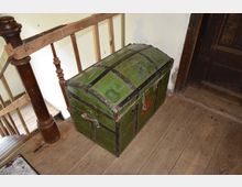 Grüne, gewölbte Holztruhe mit Metallbeschlägen und Tragegriffen, platziert auf einem Holzfußboden neben einem hölzernen Treppengeländer und einer dunklen, abgenutzten Tür.