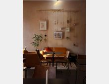 Ein gemütlicher Innenraum eines Cafés mit einer orangefarbenen Couch und dekorativen Kissen, Holztischen und -stühlen. An der Wand hängen kleine Bilder und Pflanzen an goldenen Stangen, die von der Decke herabhängen, ergänzt durch eine dezente Beleuchtung.