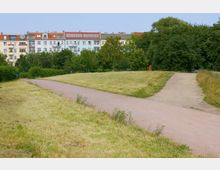 Ein Park mit einem gepflasterten und einem unbefestigten Weg, umgeben von kurz geschnittenem Gras und Bäumen. Im Hintergrund stehen mehrstöckige Wohnhäuser mit bunten Fassaden und roten Dächern.