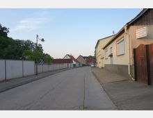 Straßenszene in einem ruhigen Wohngebiet mit gepflastertem Gehweg und schmaler Asphaltstraße, umgeben von niedrigen Wohnhäusern mit roten und beigen Fassaden. Eine weiße Mauer sowie grüne Bäume befinden sich auf der linken Seite, während ein Holztor und Blumen an Fenstern auf der rechten Seite zu sehen sind.