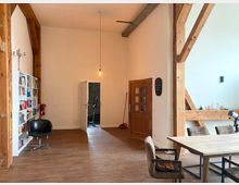 Heller, offener Raum mit Holzbalken und Holzboden; links eine Bücherwand mit Regalen und ein schwarzer Stuhl, rechts ein Esstisch mit Stühlen. Im Hintergrund sind zwei Türen, eine führt in einen weiteren Raum.