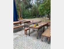 Holztische und Bänke stehen auf einer gepflasterten Fläche in einem Außenbereich, der von hohen Bäumen umgeben ist. Im Hintergrund befindet sich ein Spielplatz mit Sand, bunten Spielzeugen und Klettergerüsten.