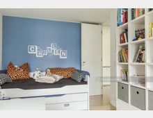 Ein Kinderzimmer mit einer blauen Wand, auf der das Wort "CHILLEN" in weißen Buchstaben steht. Vor der Wand steht ein weißes Bett mit Kissen und Stofftieren, rechts daneben ein weißes Regal mit Büchern und Aufbewahrungsboxen.