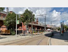 Straßenansicht einer Bahnhofsüberführung aus rotem Backstein mit mehreren Rundbögen, Oberleitungen und Gleisen für Straßenbahnen. Im Vordergrund sind Sitzplätze eines Cafés, Pflanzenkübel und Fußwege unter Bäumen zu sehen, im Hintergrund weitere Gebäude und ein Marktstand.