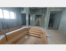 Innenansicht eines verlassenen Badebereichs mit leerem Becken, beige gefliesten Stufen und grünlichen Wandfliesen. Ein großes Fenster und ein Heizkörper befinden sich auf der linken Seite, während an der Rückwand Nischen und Durchgänge zu sehen sind.