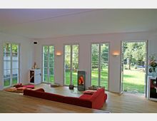 Wohnzimmer mit großen bodentiefen Fenstern und einer Tür, die zu einem Garten mit Rasen und Bäumen führt. Der Raum hat Parkettboden, ein rotes Sofa, einen Kaminofen in der Mitte und minimalistische Dekoration.