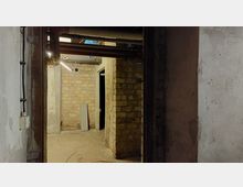 Kellerraum mit unverputzten Ziegelwänden und sichtbaren Rohren an der Decke. Im Hintergrund sind ein Türrahmen und ein beleuchtetes Leuchtstoffröhren-Licht zu erkennen.