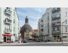Straßenszene mit Wohnhäusern und einer Kirche im Hintergrund; die Kirche hat eine große, dunkle Kuppel und einen markanten Kirchturm. Links befindet sich ein kleines Geschäft mit roter Markise, und auf der Straße stehen parkende Autos.