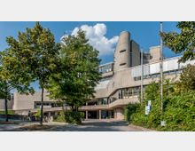 Außenansicht eines modernen Gebäudes aus Beton mit geschwungener Architektur, großen Fenstern und einem hohen Turmelement. Umgeben von Bäumen und Grünflächen, mit einem Gehweg im Vordergrund.