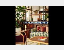 Wohnzimmer mit modernem, eklektischem Design: Ein Sofa mit geometrischem Muster, bunte Kissen, ein Tisch mit einer Zeitschrift, Holzmöbel und üppige Pflanzen. Im Hintergrund sind Regale mit Dekorationen und eine Wand mit weißen Fliesen und blauem Muster zu sehen.