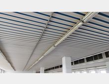 Industriehalle mit einer Metall-Deckenkonstruktion, bestehend aus weißen Paneelen mit blauen Linien. An der Decke sind lange Leuchtstofflampen befestigt, und unten sind weiße Säulen und Fenster zu sehen.