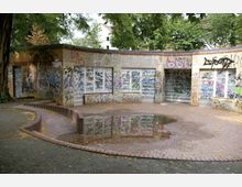 Halbrundes, niedriges Gebäude aus gelben Backsteinen, dessen Wände großflächig mit Graffiti bedeckt sind; davor eine gepflasterte Fläche mit einer Pfütze und umliegenden Bäumen.