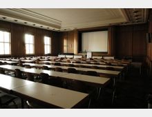 Ein Konferenzraum mit Holzwänden und mehreren Reihen von Tischen und Stühlen, die auf eine Bühne mit einem Rednerpult und einer weißen Leinwand ausgerichtet sind. Tageslicht fällt durch große, rechteckige Fenster an der linken Seite des Raums.