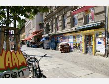 Straßenszene in einer Stadt mit historischen Gebäuden; an den Fassaden befinden sich bunte Plakate und Graffiti. Im Vordergrund stehen Fahrräder, ein kleiner Markt mit Obst- und Gemüsekisten ist zu sehen, teilweise unter farbigen Markisen und einem blauen Sonnenschirm.