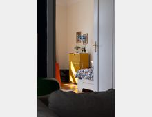 Blick in ein gemütliches Zimmer mit einer offenen Tür. Im Raum stehen ein gelber Schrank, ein Bett mit floral gemusterter Bettwäsche und eine Vase mit blauen Orchideen; die Wand ist cremefarben, und Sonnenlicht fällt durch ein Fenster herein.