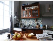 Eine Küche mit einem Mix aus traditionellem und modernem Design. Eine Holzoberbauvitrine mit Glastüren hängt über einer Arbeitsfläche mit Spüle und buntem Fliesenmuster, daneben ein moderner Dunstabzug. Im Vordergrund steht ein Holztisch mit Körben voller Äpfel und Zeitungen.