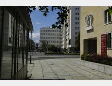 Straßenszene mit einem gepflasterten Gehweg, flankiert von Glasfenstern auf der linken Seite und einem beigefarbenen Gebäude auf der rechten Seite mit einem steinernen Adlerrelief und einer roten Informationstafel. Im Hintergrund sind moderne Wohngebäude und ein Hotel zu sehen.