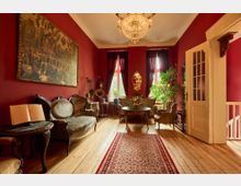 Wohnzimmer im klassischen Stil mit roten Wänden, historischen Möbeln und Holzboden. Ein Kronleuchter hängt von der Decke, an den Fenstern hängen schwere Vorhänge, und ein großer Wandteppich sowie zahlreiche Pflanzen und dekorative Objekte vervollständigen das elegante Ambiente.