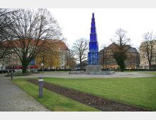 Öffentlicher Platz mit einer hohen, rechteckigen Skulptur aus blauem Glas auf einem Betonsockel, umgeben von gepflasterten Wegen und Rasenflächen. Im Hintergrund stehen Gebäude mit roten Dächern und großen Bäumen ohne Blätter.