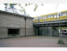 Ansicht eines Bahnhofs mit einem gelben Zug der Hochbahn, der über eine Brücke fährt. Im Vordergrund ist ein Platz mit Pflastersteinen, ein Laternenpfahl und ein modernes Gebäude mit Glasfassade sowie ein älteres gemauertes Gebäude zu sehen.