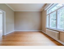 Unmöblierter Wohnraum mit hellem Holzdielenboden, beigefarbenen Wänden und weißer Decken- sowie Fußleisten. Rechts große, mehrteilige Fensterfront mit Blick ins Grüne und darunter ein weißer Heizkörper; links eine offene Türöffnung zu einem Nebenraum.