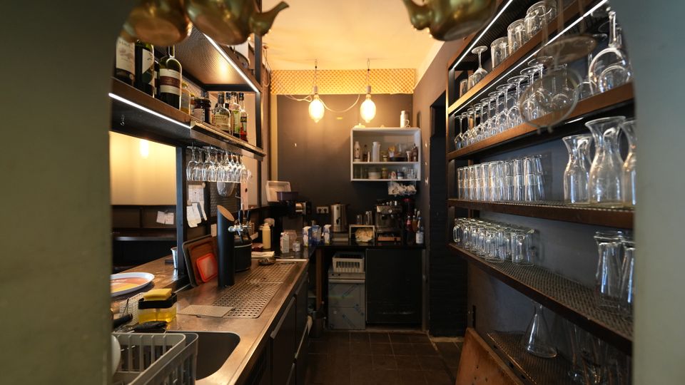 Blick in einen schmalen Bar- und Spülbereich mit Edelstahl-Arbeitsfläche, Spülbecken und Regalen mit Flaschen und hängenden Gläsern. Rechts stehen mehrere Holzregale voller Trinkgläser, im Hintergrund sind eine Kaffeemaschine und weitere Barutensilien unter warmen Deckenleuchten zu sehen.