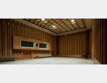 Großer rechteckiger Raum mit hohen Wänden, die mit vertikalen Holzpaneelen verkleidet sind. Die Decke hat ein geometrisches Muster, und der Raum enthält Sitzbänke entlang der Wände, eine Wanduhr und in der Ecke einen schwarzen Flügel.