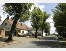 Wohnstraße in einer ländlichen Umgebung mit einem Einfamilienhaus mit roten Ziegeldächern, Holzfenstern und einem Vorgarten, umgeben von Bäumen. Links neben dem Haus ist eine Mauer mit einem Tor, rechts führt die Straße weiter durch von Bäumen gesäumte Umgebung.