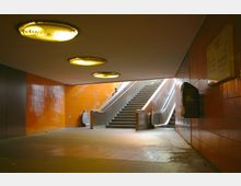Unterführung mit orangefarbenen Fliesenwänden, beleuchtet durch runde Deckenlampen. In der Mitte befinden sich eine Treppe und eine Rolltreppe, die nach oben führen.