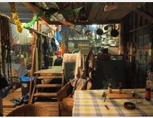 Ein rustikaler Innenraum einer Scheune oder Hütte mit Holzboden und Holzstruktur, dekoriert mit bunten Girlanden. Im Vordergrund steht ein Tisch mit kariertem Tischtuch, Korbstühlen und verstreuten Gegenständen, im Hintergrund befinden sich Regale mit Werkzeugen, Küchenutensilien und Kochgeschirr.