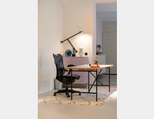 Ein minimalistisches Homeoffice mit einem Schreibtisch aus Holz und Metallrahmen, einem ergonomischen Bürostuhl und einer Schreibtischlampe. Im Hintergrund sind eine pastellfarbene Kommode, warme cremefarbene Wände und eine Türöffnung zu sehen, ergänzt durch einen hellen Teppich auf dem Boden.