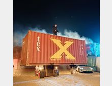 Ein großer, rotbrauner Schiffscontainer mit einem gelben "X" steht in einer nächtlichen Umgebung auf einem Gabelstapler. Auf dem Container steht eine Person, umgeben von aufsteigendem Rauch, während im Hintergrund ein Sportwagen und weitere Container zu sehen sind.
