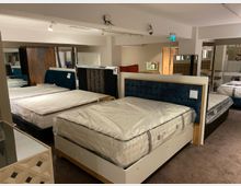 Ausstellungsraum eines Möbelgeschäfts mit mehreren ausgestellten Betten, die Matratzen sind in Plastik verpackt. Im Hintergrund sind gespiegelte Wände, Schränke und Kopfteile in verschiedenen Designs sichtbar.