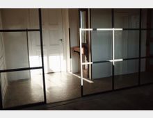 Raum mit einer weißen Holztür im Hintergrund, geteilt durch eine Glaswand mit schwarzen Metallstreben; Sonnenlicht fällt herein und erzeugt Schattenmuster auf dem Boden. Rechts steht ein an der Wand befestigter kleiner Holztisch.