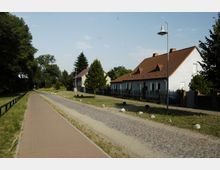 Ländliche Straße mit Kopfsteinpflaster, flankiert von einem gepflasterten Gehweg und einfachen Einfamilienhäusern mit roten Ziegeldächern auf der rechten Seite. Auf der linken Seite erstreckt sich eine Grünfläche mit Bäumen, beleuchtet von vereinzelten Straßenlaternen.