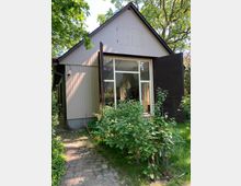 Ein kleines Gebäude mit Satteldach und vertikaler Holzverkleidung, dessen Vorderseite eine große verglaste Tür mit geöffneten schwarzen Holzläden hat. Der Eingang ist von Sträuchern und einem gepflasterten Weg umgeben, im Hintergrund sind Bäume zu sehen.