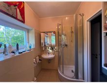 Kleines Badezimmer mit beige gefliesten Wänden, einem Waschbecken mit Spiegel und einer halbrunden Eckdusche mit Glasabtrennung. Links sorgt ein großes Fenster über einer Ablage mit Pflegeprodukten für Tageslicht und Blick ins Grüne.