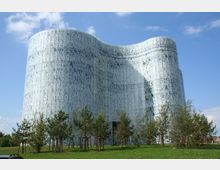 Ein modernes Gebäude mit wellenförmiger Glasfassade, die mit abstrakten Mustern gestaltet ist, umgeben von einer Grünfläche mit jungen Bäumen. Das Gebäude steht unter einem blauen Himmel mit vereinzelten Wolken.