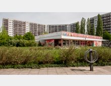 Flaches Gebäude mit bemalter Fassade und großen Fenstern, umgeben von Sträuchern und einer Wiese, im Hintergrund stehen mehrere hohe Wohnblöcke mit Balkonen. Im Vordergrund befindet sich ein Fahrradbügel auf einem Pflasterweg.