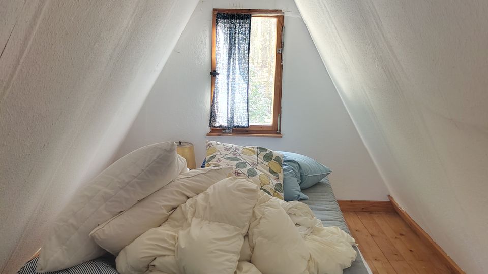 Kleines Schlafzimmer im Dachboden mit stark geneigten, weiß verkleideten Wänden und sichtbarem Holzbalken am First. Ein niedriges Bett steht auf Holzdielen vor einem kleinen Fenster mit Holzrahmen und Vorhang.