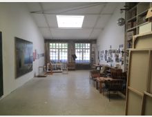 Ein geräumiges Künstleratelier mit hoher Decke, weiß gestrichenen Wänden und einem großen Fenster mit Gitterrahmen im hinteren Bereich. Im Raum stehen Staffeleien, gerahmte Kunstwerke, Sitzmöbel und Regale, an den Wänden hängen Gemälde und Fotografien.