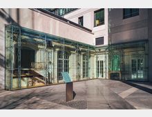 Innenhof mit einer historischen Wandverkleidung, geschützt durch eine moderne Glaskonstruktion mit Metallrahmen. Der Boden ist mit Pflastersteinen ausgelegt, und eine Informationstafel steht im Vordergrund.